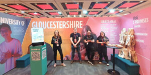 UoG Exhibition stand with large bright photography of students and stand exhibitors stood infront