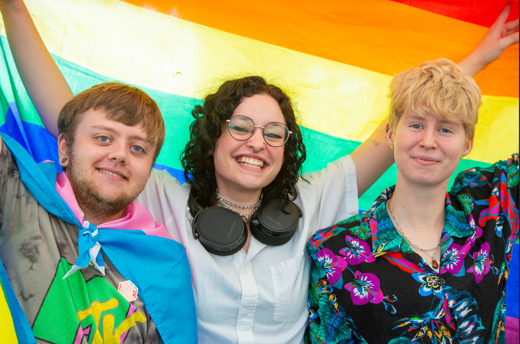 3 students holding a rainbow flag behind them.